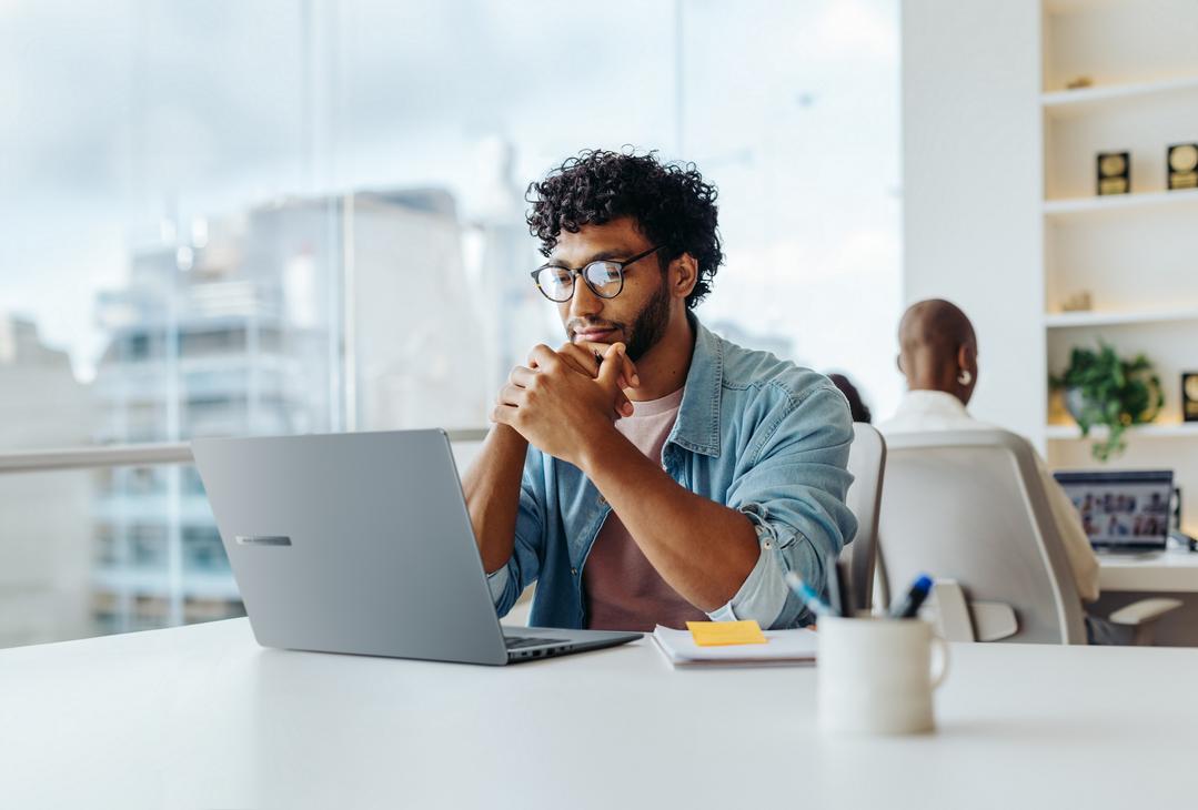 Homem usando notebook em escritório moderno, sentado à mesa com postura atenta, representando boas práticas de ergonomia no trabalho.