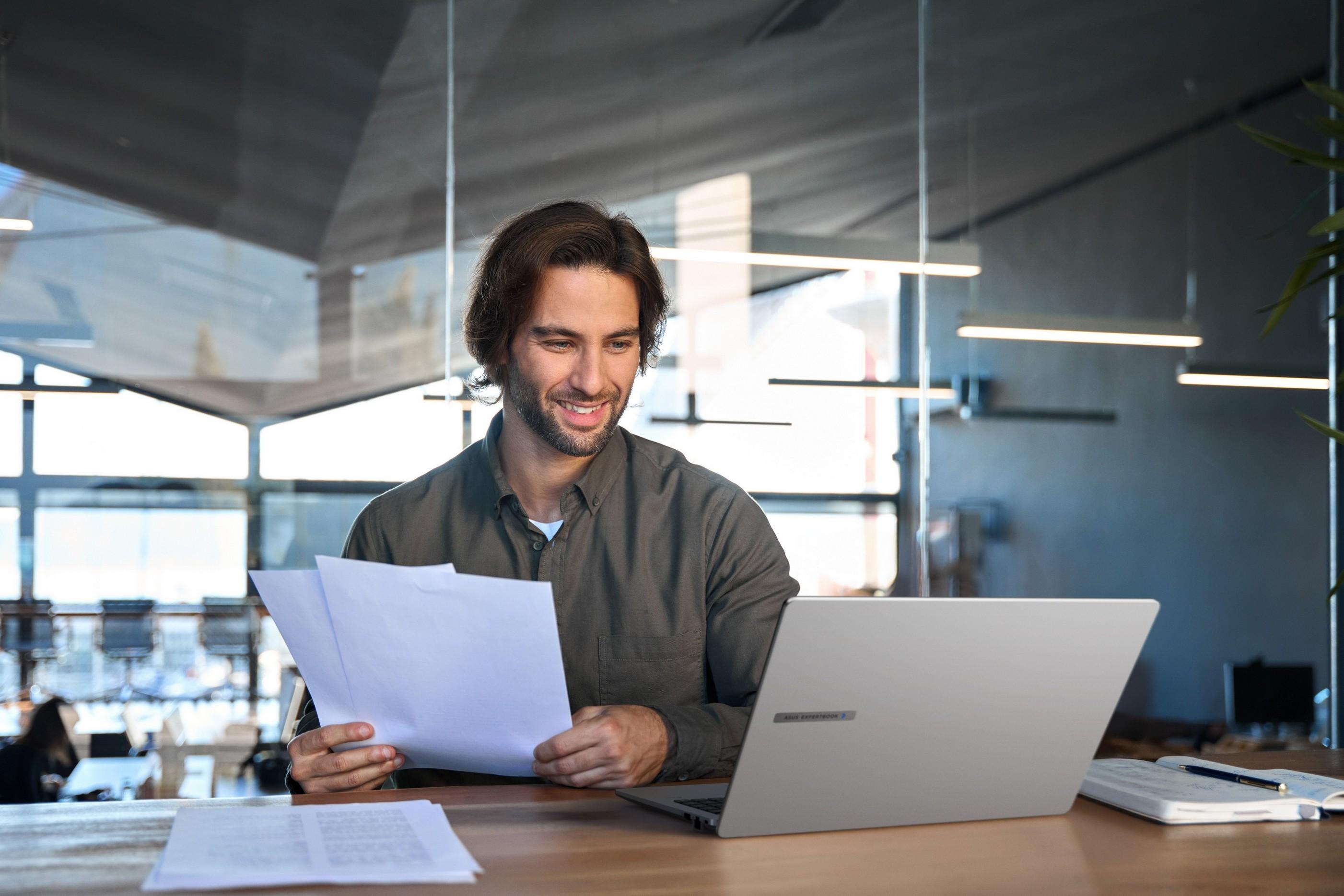 Homem trabalhando em notebook com postura correta e documentos em mãos, representando práticas de ergonomia no trabalho em escritório moderno.