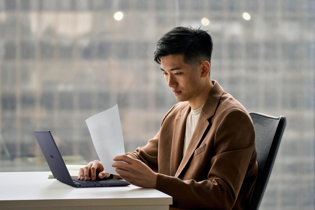 Homem sentado em frente a notebook segurando documentos, ilustrando ergonomia no trabalho com postura adequada em ambiente corporativo.